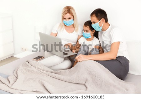Family in medical face masks lying on a bed with a laptop. Concept of watching video, working together and quarantine during a COVID-19 coronavirus epidemic