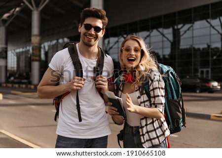 Surprised blonde woman and happy brunette man in sunglasses poses near airport. Cheerful travelers smiling and holding backpacks.