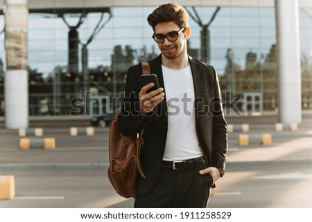 Cool brunette man in eyeglasses chats in phone. Happy guy in black suit holds brown backpack and smiles outside.