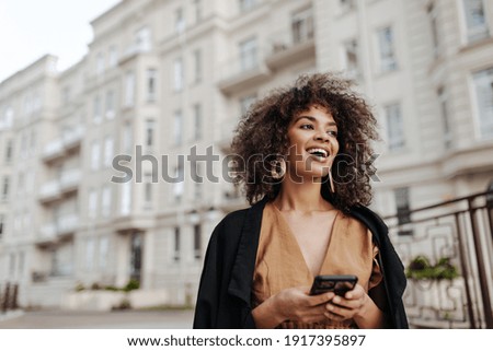 Cheerful African woman holds smartphone. Curly brunette lady in brown blouse and black coat smiles and walks outdoors.