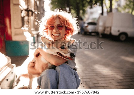 Happy curly woman in jeans smiles sincerely and hugs corgi. Good-humored girl in red sunglasses plays with dog on street.