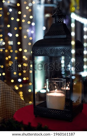 an iron lantern with glass candles and a flame with glowing warm light on a night terrace with bokeh from festive garlands on the window in defocus.