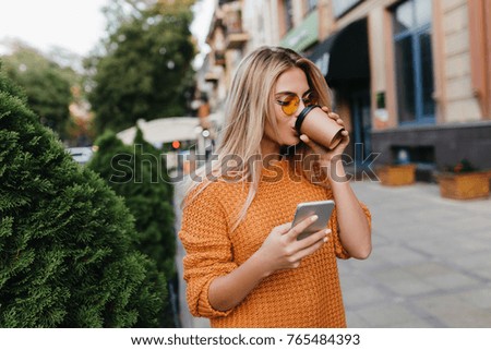 Encantadora joven rubia esperando un mensaje de teléfono mientras toma café en la calle. Chica elegante con suéter amarillo posando en un callejón sosteniendo el smartphone y disfrutando del cappuccino.