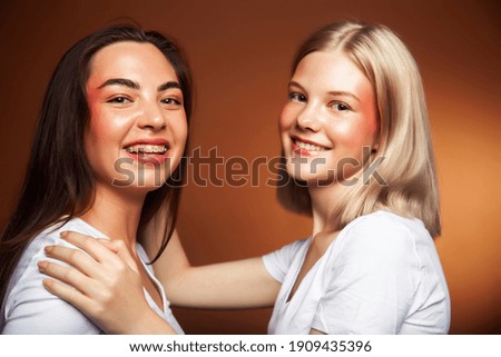two pretty diverse girls happy posing together: blond and brunette, caucasian and asian on brown background, lifestyle people concept