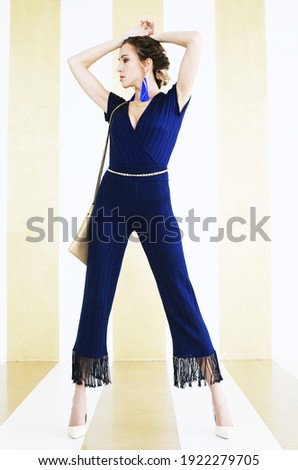 Full-length portrait of beautiful fashionable young woman in blue overalls, against the background of a striped wall, fashion concept