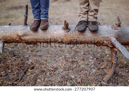 man and woman in trousers and boots standing on a fallen tree in a pine forest