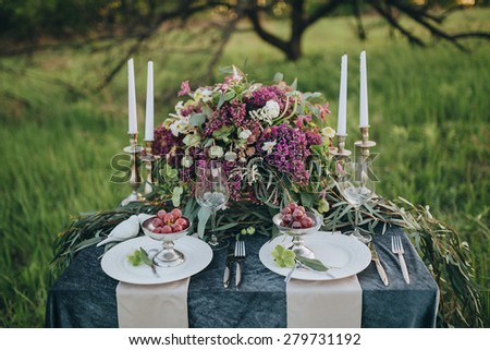decorated table for two with a dark cloth decorated with floral composition of lilac and green candles on a background of grass and orchard