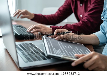 Asian woman programmer holding code data on clipboard and discussion with partner about new project while working together to writing code for new website or application in software development office
