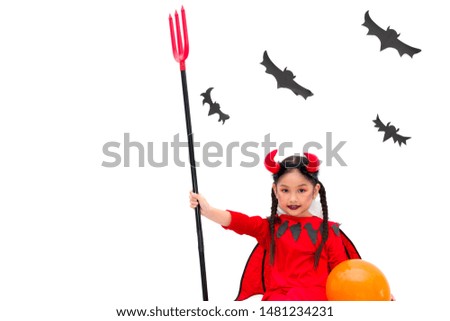 Asian cute little girl is wearing red Devil costume and holding trident while sitting leaning against the wall. At the white backdrop have a black bats made of cardboard. Halloween festival concept.