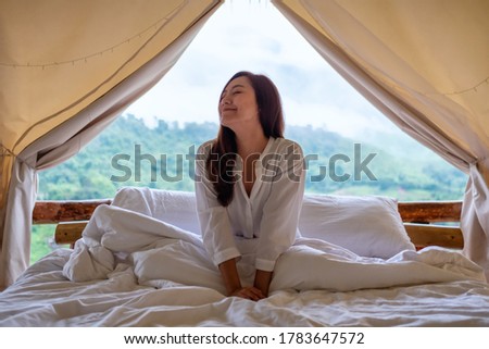 Imagen de una joven sentada en una cama blanca por la mañana con una hermosa vista de la naturaleza fuera de la carpa