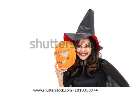 Asian woman wearing Halloween costume as witch in black cloak, on white background, holding orange pumpkin mask, looking at camera