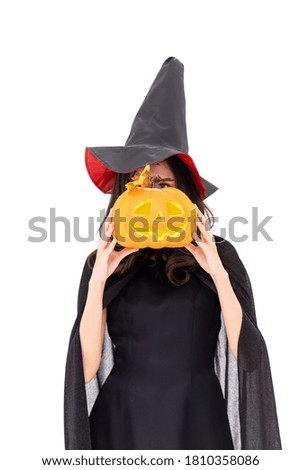 Asian woman wearing Halloween costume as witch in black cloak, on white background, cover face with orange pumpkin Jack O Lantern, looking at camera