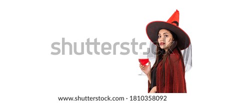 Asian woman wearing Halloween costume as witch in red cloak, on white background, holding red wine glass, looking at camera