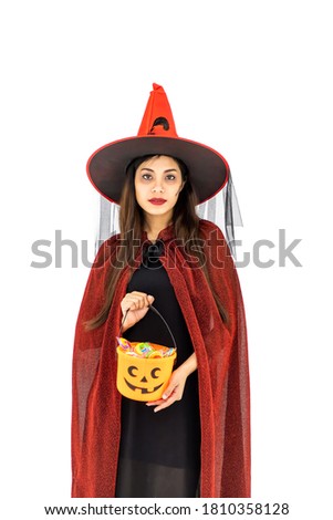 Asian woman wearing Halloween costume as witch in red cloak, on white background, holding orange candy pumpkin bucket for trick or treat, looking at camera