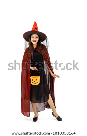Asian woman wearing Halloween costume as witch in red cloak, on white background, holding orange candy pumpkin bucket for trick or treat, looking at camera