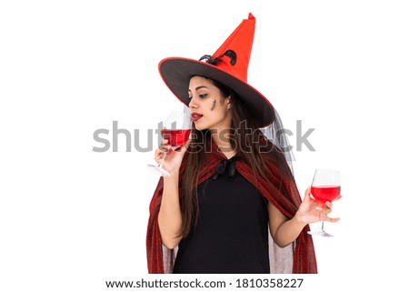 Asian woman wearing Halloween costume as witch in red cloak, on white background, holding glass and drinking red wine
