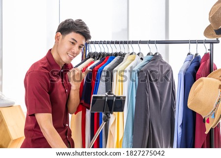 Young Asian male online merchant blogger using mobile phone camera with selfie stick to live video to review and sell product. Social media, Influencer, and online shopping concept.