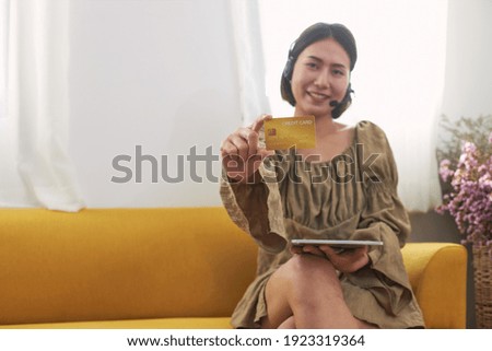 A happy Asian woman sitting on a yellow sofa, holding a credit card to the front. Expresses the concept of buying products online. Cashless society ideas.