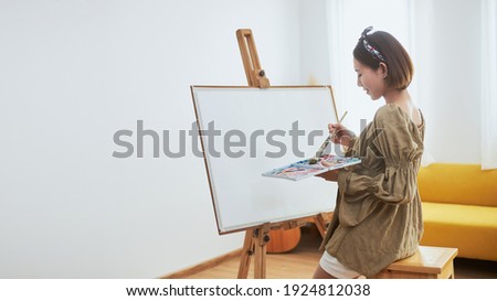 An Asian woman who loves art or a female artist is holding a paint palette and paintbrushes while happily painting on white paper at home.