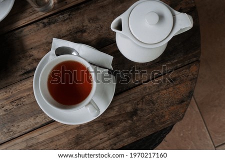 A top view of a cup of tea on a saucer with napkin and spoon on a wooden table with teapot