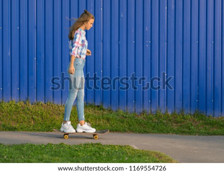 Slender trendy young woman skateboarding in town along a path in front of a colorful blue wall in evening light