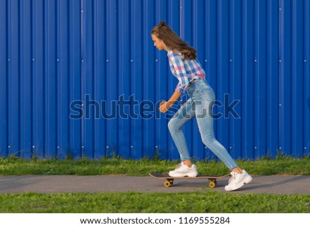 Trendy slender young woman with long brown hair skateboarding at dusk along an urban path in front of a blue wall