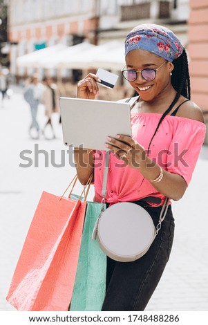 Happy young african american woman make shopping in the tablet with credit card and holding shopping bags on the street