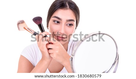 beautiful young smiling woman holding makeup brush and looking her face in the mirror. isolated on white background.