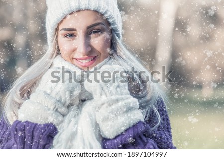 Portrait of happy woman in warm clothing on snowing winter day outdoors. Snowflakes falling on cheerful girl wearing wool cap, scarf and purple sweater. Snow and happiness concept