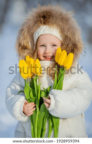 cute little girl in white winter clothes and a knitted hat walks in the winter forest with tulips. early spring, international women's day