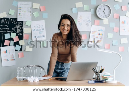 Cute, smiling business girl with glasses in the office leaning on the desk. On a gray background pasted stickers, graphics, work plans.