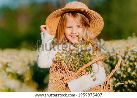 Cute little blonde girl in a cotton dress and straw hat walks in a field of daisies collects them in the basket
