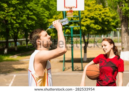 Young Athletic Couple Taking a Refreshing Break on Basketball Court, Woman Looking On As Man Empties Water Bottle Onto Face