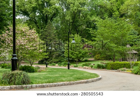 Nature walk pathway. Path down curved road. Black street steel lamps on manicured landscaping.Shrub and trees down street. Secret garden walkway.