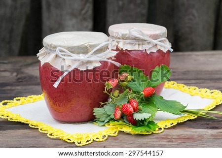 Fresh homemade puree of wild strawberry with sugar in banks and bouquet of strawberries on a napkin with yellow edging. The rustic style. Selective focus
