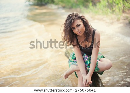 Beautiful brunette woman in bright clothes sits on the bank of the river on a background of water