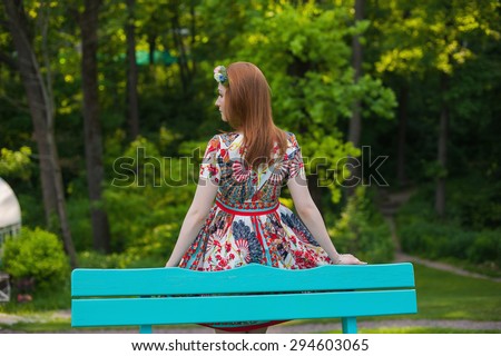 Beautiful young red-haired woman in a bright dress stands back near the blue benches in the garden