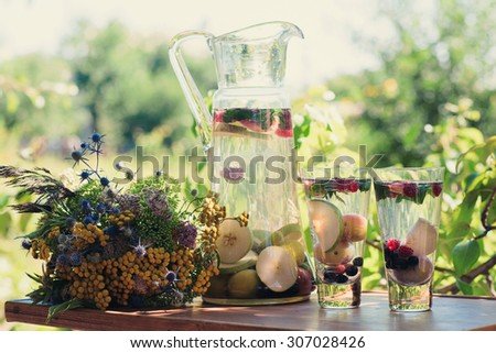 Summer cocktails with seasonal fruit in transparent glass carafe stands on a table in a garden next to a glass beaker with a drink of vegetarian food