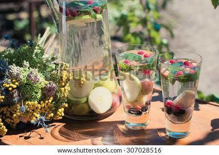 Summer cocktails with seasonal fruit in transparent glass carafe stands on a table in a garden next to a glass beaker with a drink of vegetarian food