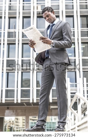 Black businessman reading newspaper outdoor. Behind building with windows