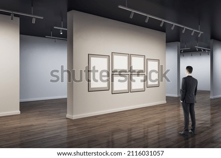Businessman in modern concrete exhibition hall interior looking at blank white mock up posters on wall and wooden flooring with reflections. Art and museum concept