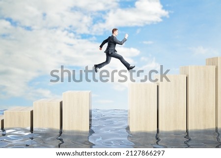 Businessman jumping over obstacles between wooden chart bars above water on bright blue sky background. Challenge and success concept