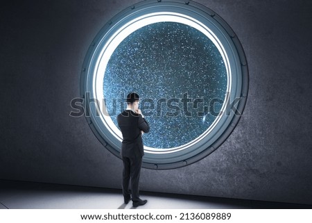 Back view of young business man looking out of round illuminator with starry sky cosmos view in concrete interior. Future and creativity concept