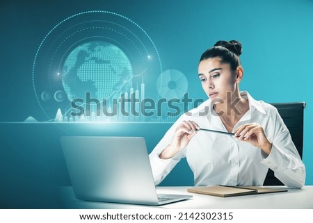 Attractive european businesswoman working at desktop with laptop and abstract glowing digital business chart and globe on blurry blue background. Trend, finance and economy concept. Double exposure