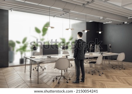 Thoughtful young man standing in modern coworking office interior with equipment, furniture, decorative plants and matte glass partition. Workplace concept
