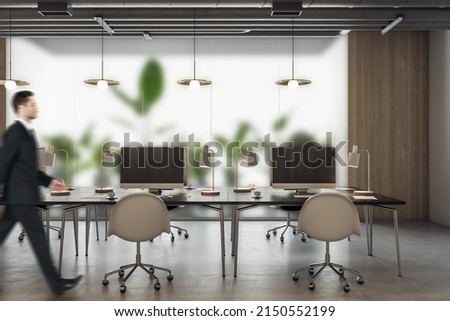Thoughtful young businessman walking in modern coworking office interior with equipment, furniture, decorative plants and matte glass partition. Workplace concept