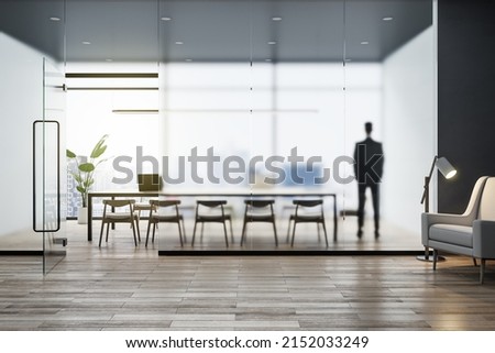 Attractive young european businessman standing in modern concrete office interior with wooden flooring, matte partition glass, furniture, equipment and city view with daylight. Worker and ceo concept