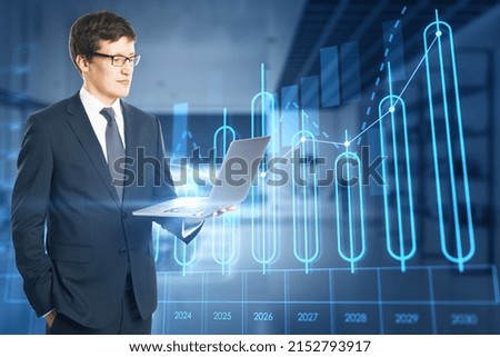 Attractive young european businessman using laptop with growing annual business chart report hologram on blurry office interior background. Analysis and company concept. Double exposure