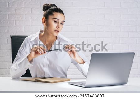 Business process and job concept with pretty young woman sitting at white desk, looking at laptop screen and carrying pencil in hands on light brick wall background