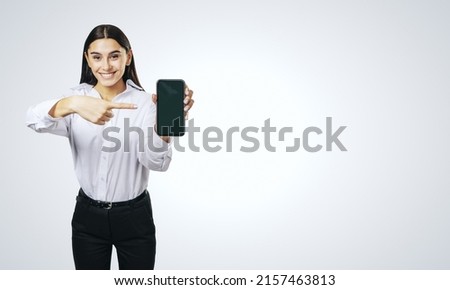 Mobile application concept with happy girl in white shirt showing modern cellphone with blank screen on abstract light grey background with place for your logo or text, mockup